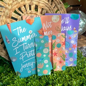 May include: A collection of colorful bookmarks and a book titled "The Summer I Turned Pretty" by Jenny Han. The bookmarks feature seashell and aquatic designs in shades of blue, green, orange, and purple. The book has a turquoise cover.
