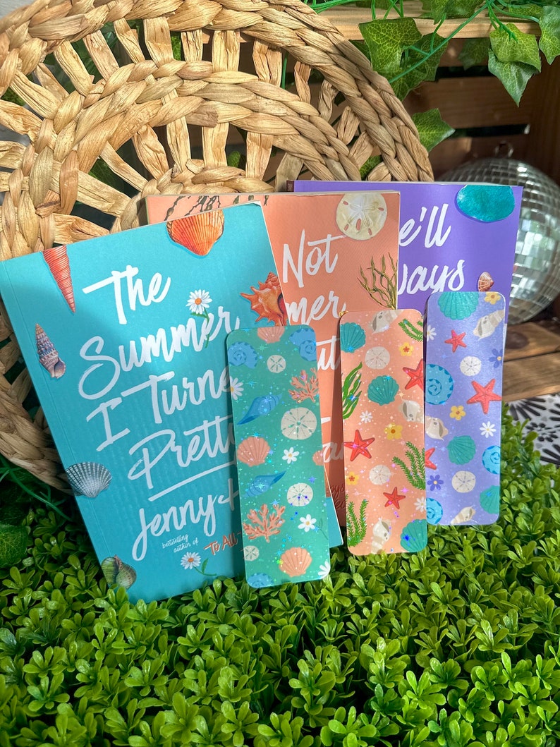 May include: A collection of colorful bookmarks and a book titled "The Summer I Turned Pretty" by Jenny Han. The bookmarks feature seashell and aquatic designs in shades of blue, green, orange, and purple. The book has a turquoise cover.