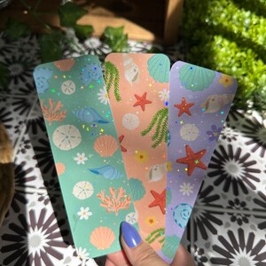 May include: Three colorful bookmarks with a sea life theme. Each bookmark features a different background color: mint green, peach, and lavender. The designs include seashells, starfish, coral, and daisies. The bookmarks have a holographic glitter effect.