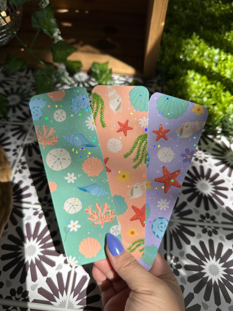 May include: Three colorful bookmarks with a sea life theme. Each bookmark features a different background color: mint green, peach, and lavender. The designs include seashells, starfish, coral, and daisies. The bookmarks have a holographic glitter effect.