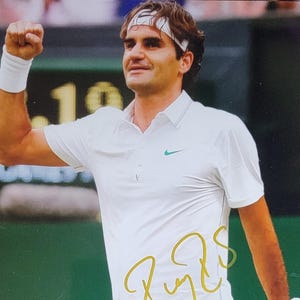 May include: A signed photograph of a tennis player wearing a white polo shirt with a green Nike logo, white wristband, and headband. The player is holding a tennis racket and celebrating with a raised fist. The signature is in gold.