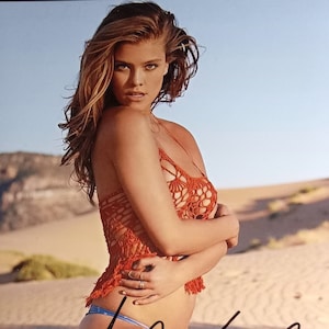 Peut inclure: Une femme portant un haut en crochet rouge et un bas de bikini bleu pose dans un décor désertique. Elle tient ses bras autour de son torse et ses cheveux flottent au vent. L'image est signée "Lily Aldridge".