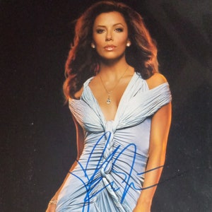 Puede incluir: Una mujer con un vestido azul claro con un diseño anudado. Lleva un collar plateado y tiene el pelo peinado en ondas sueltas. El fondo es negro.