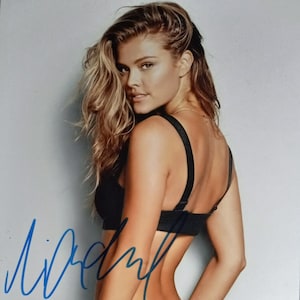 Peut inclure: Une femme aux longs cheveux blonds portant un soutien-gorge noir et des sous-vêtements noirs avec une boucle. La femme regarde par-dessus son épaule et a une signature écrite à l'encre bleue.