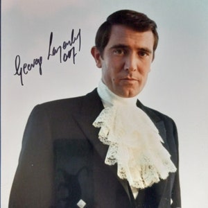 Może przedstawiać: Podpisany portret George'a Lazenby'ego z podpisem "George Lazenby 007". Ma na sobie czarny smoking z białą koronkową koszulą. Zdjęcie pochodzi z filmu "W tajnej służbie Jej Królewskiej Mości". Widoczna jest naklejka autentykacyjna PSA.
