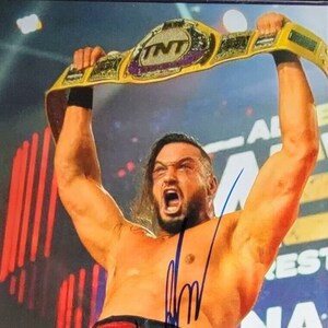 May include: A professional wrestler holding up a gold and purple TNT championship belt. The wrestler is shirtless and has a signature on the image. The background has the words "All Elite Wrestling" and "Dynamite".