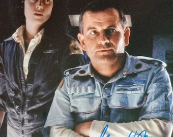 May include: A black and white photo of two people, a woman with dark hair and a man with short brown hair. The man is wearing a blue denim shirt and has his arms crossed. The woman is wearing a dark blue jacket. The man's signature is visible in blue ink.
