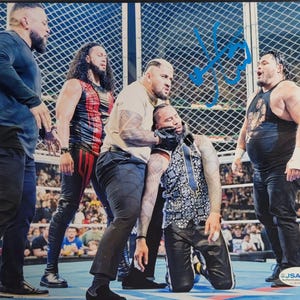 May include: A signed photograph featuring five wrestlers in a ring with a chain-link fence background. One wrestler kneels while others stand around, wearing black pants, tank tops, and shirts. A blue autograph is visible on the upper right side.