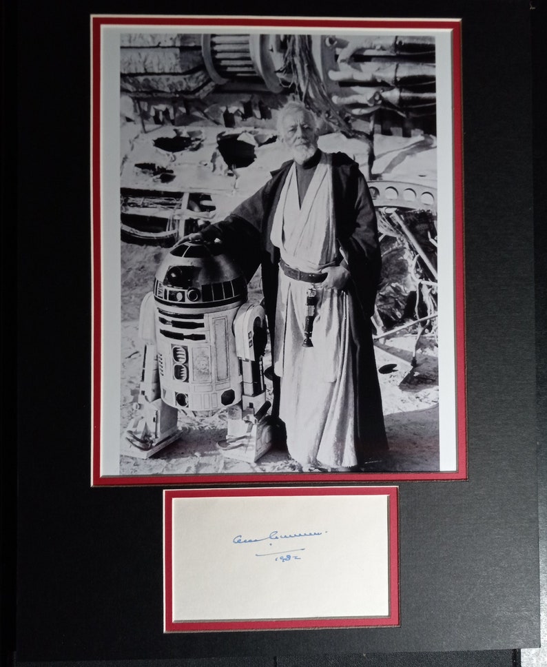 May include: Black and white photo of a man in a white robe standing next to a robot. The man is holding a lightsaber and the robot is R2-D2. The man's signature is in blue ink below the photo. The signature reads "Alec Guinness 1994."