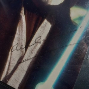 May include: A black and white photograph of a person holding a lightsaber. The person's signature is written in black ink on the photo. The signature reads "Mark Hamill".
