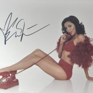 Puede incluir: Una mujer en lencería roja y un boa de plumas posa con un teléfono rojo de disco. El teléfono está en el suelo y su pie está apoyado en él. La mujer mira a la cámara y sonríe. La firma "Kristy" está escrita en tinta negra en la esquina superior izquierda de la imagen.