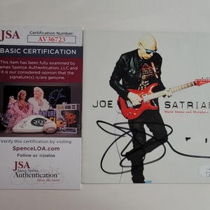 May include: An autographed Joe Satriani album cover featuring a red electric guitar and the album title "Black Swans and Wormhole Wizards." Includes a JSA authentication certificate with the number AV36723.