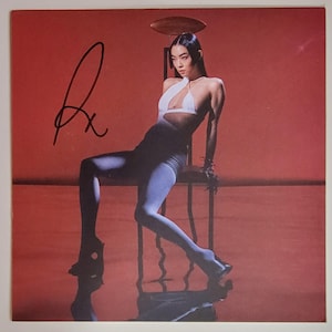 May include: A signed album cover showing a person in a white halter top, dark leggings, and blue tights, seated on a chair against a red background. The person is leaning forward, with a reflective surface below.