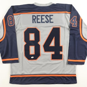 May include: A gray hockey jersey with navy blue sleeves and orange accents. The jersey features the name "REESE" across the back and the number "84" in navy blue. The number "8" is on the left sleeve and "4" on the right sleeve.