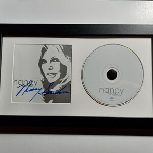 Peut inclure: Un présentoir encadré noir avec un passe-partout blanc. Le présentoir présente une photo en noir et blanc de Nancy Sinatra avec sa signature en encre bleue. Un CD avec le texte "Nancy Sinatra" est également présenté.