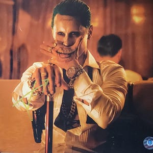 May include: A portrait of a person with green hair and a painted smile, wearing a white shirt, gold watch, and multiple rings. The person is holding a cane and has a tattoo on their wrist. The background is blurred with warm tones.