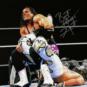 Puede incluir: Una foto en blanco y negro de dos luchadores en un ring de lucha libre. El luchador de arriba lleva pantalones negros, una camiseta negra y morada y botas negras. El luchador de abajo lleva pantalones blancos con un diseño morado y dorado y botas blancas. El luchador de arriba tiene su brazo alrededor del cuello del luchador de abajo. El luchador de arriba mira a la cámara con una expresión decidida. El luchador de abajo mira hacia abajo al tapete. La foto está firmada "Bret Hart..." con tinta plateada.