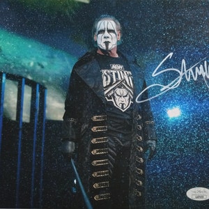 May include: A black and white photo of a wrestler wearing a black coat with gold buttons and a black shirt with a white skull and crossbones logo. The wrestler has a white face paint design and is holding a black stick. The wrestler's signature is in the top right corner of the photo.