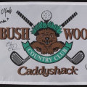 May include: A white flag with a brown and green logo for Bushwood Country Club. The logo features a cartoon bear with crossed golf clubs and a golf ball. The flag is signed by Michael O'Keefe, Bill Murray, and Chevy Chase.
