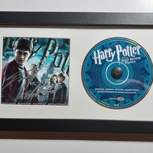 Puede incluir: Una presentación enmarcada que incluye un CD y una foto de la película "Harry Potter y el misterio del príncipe". El CD presenta un diseño azul y plateado con el título de la película y las palabras "Banda sonora original de la película". La foto muestra a Daniel Radcliffe como Harry Potter, con otros personajes en el fondo.