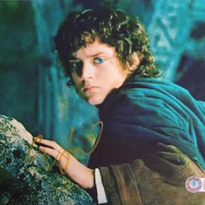 Può includere: Una fotografia a colori di un giovane con capelli castani ricci e occhi blu, che indossa un mantello con cappuccio verde acqua scuro. L'immagine ha una firma con inchiostro dorato e un piccolo adesivo nell'angolo in basso a destra. Lo sfondo è sfocato, verde acqua scuro.