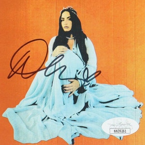 May include: A woman with long dark hair sits on an orange background wearing a light blue gown and a crown. She is holding her hands in her lap. The image is signed "Demi".