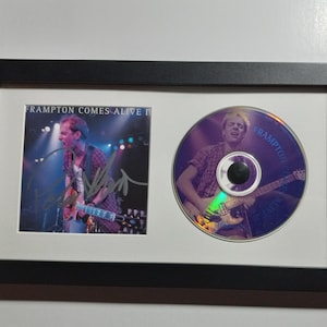 May include: Framed CD and photo featuring Peter Frampton. The photo and CD are in a black frame with a white mount. The photo shows Frampton playing guitar on stage. The CD has the album title "Frampton Comes Alive!"