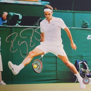 May include: A signed photograph of a tennis player in action, mid-swing. The player is wearing a white athletic outfit, headband, and shoes. A tennis racket and ball are visible. The background includes a green court and spectators.