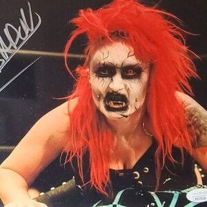 May include: A woman with bright red hair and heavy makeup, including black eyeliner and dark lipstick, is wearing a black top and has a tattoo on her right arm. The image is signed "ABRPKL".