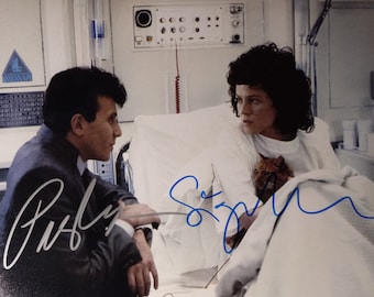 May include: A black and white photo of two actors, Sigourney Weaver and  a man, in a hospital room. The photo is signed by both actors. Sigourney Weaver is holding a cat in her arms. The photo is a signed 8x10 print.