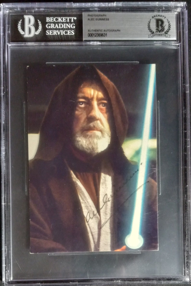 May include: A black and white photograph of Alec Guinness as Obi-Wan Kenobi from Star Wars. The image is signed in black ink. The photograph is encased in a Beckett Grading Services holder with the number 0001236931.