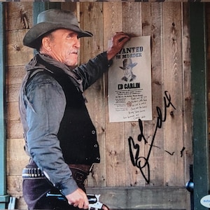 May include: A man in a cowboy hat and vest stands next to a wooden wall, holding a wanted poster that reads "Wanted for Murder" with the name "Ed Carlin." The poster is signed. He is also holding a gun.