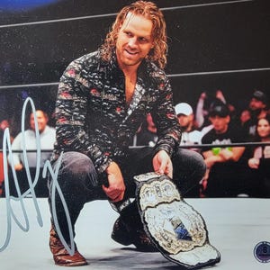 May include: An autographed photo of a smiling man in a black patterned shirt and jeans, kneeling on a wrestling ring. He holds a championship belt with a silver and blue design. The background shows a crowd of people.