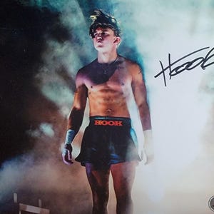 May include: An image of a person in black shorts with the word "HOOK" in red. The person is standing in a smoky environment with a signature in the upper right corner. The person is wearing wristbands.