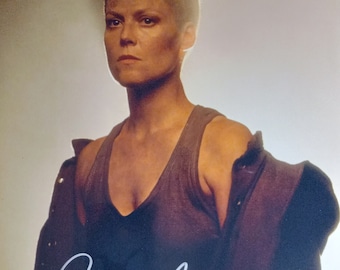 May include: A black and white photo of a woman with short, shaved hair. She is wearing a dark tank top and a jacket. The photo is signed "Sigourney" in white ink.