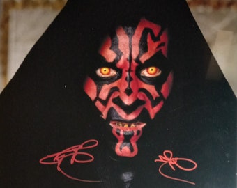 May include: A framed image featuring a close-up of a character with red and black facial markings, yellow eyes, and a black hood. The image includes two red signatures and a certification sticker. The character's face is the focal point.