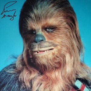 May include: A portrait of Chewbacca from Star Wars, with his characteristic fur, facial features, and bandolier. The image has a blue background and includes an autograph in the upper left corner. A Beckett Authentication sticker is visible.