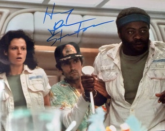 May include: A black and white photo of three people from the movie Alien. The people are standing in a room with white walls. The person on the left is a woman with dark hair. The person in the middle is a man wearing a hat and a floral shirt. The person on the right is a man with a beard and a blue headband. The photo is signed "H.R. Giger".