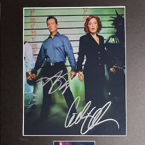 Op de afbeelding: Een zwart-witfoto van twee mensen die voor een politiebureaustandwand staan. De vrouw rechts draagt een zwart pak en de man links draagt een blauw shirt en een zwarte broek. Beide zijn gesigneerd met hun namen. De foto is ingelijst en heeft een klein beeld van het "X-Files"-logo rechtsonder.