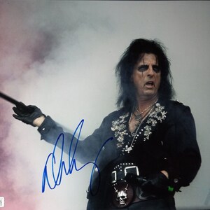 May include: A black and white photograph of a musician with long black hair and a black shirt with silver embellishments. He is holding a black and white cane and is wearing a black belt with a skull buckle. The musician is performing on stage in front of a white background with smoke in the air. The photo is signed in blue ink with the name "Alice Cooper".