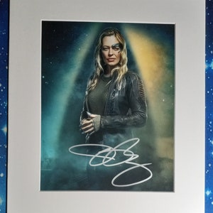 Può includere: Una foto in bianco e nero di una donna con i capelli biondi che indossa una giacca di pelle nera e una camicia verde. Ha un segno blu e nero sulla fronte. La foto è firmata con una firma bianca.