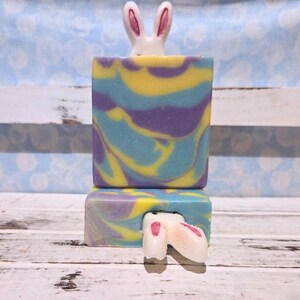 May include: Two stacked, colorful soap bars with a bunny theme. The top bar has a white bunny head with pink ears peeking out. The soap bars have a swirl pattern in yellow, blue, and purple. The bottom bar has a bunny tail.