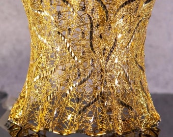 May include: A decorative gold-colored metal vase with a unique openwork design. The vase has a flared, ruffled top and a curved body. The intricate pattern creates a delicate, airy appearance, reflecting light and adding visual interest.