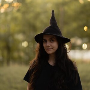 May include: A dark charcoal gray crocheted witch hat featuring a tall, pointed crown and a wide, slightly curved brim. The hat is crafted with a visible knit texture and is worn atop long, dark hair, paired with a plain black t-shirt.