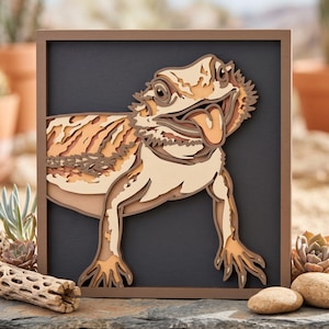 Könnte beinhalten: Ein mehrschichtiges Wandbild aus Holz, das eine Bartagame mit herausgestreckter Zunge zeigt. Die Eidechse ist in Beige-, Creme- und Brauntönen gehalten und befindet sich vor einem dunkelgrauen Hintergrund in einem quadratischen Holzrahmen.