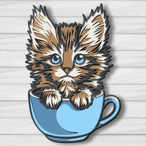 Puede incluir: Una pieza de arte en papel en capas de un gatito en una taza azul. El gatito tiene pelaje marrón y blanco, ojos azules y está sentado en una taza azul claro. El fondo es un patrón de vetas de madera blanca.