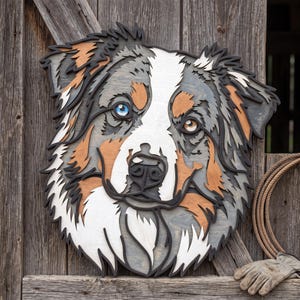 Peut inclure: Une décoration murale en bois multicouche représentant une tête de chien berger australien. Le design présente des découpes complexes dans des tons gris, blancs et fauves, avec un œil bleu vif et un œil marron, souligné par un contour noir.