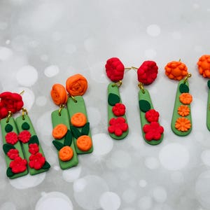 Puede incluir: Una colección de pendientes hechos a mano de arcilla polimérica. Los pendientes presentan una base verde con diseños florales rojos y naranjas. Los pendientes tienen postes dorados. Las flores incluyen rosas y otros diseños florales detallados.