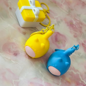 Könnte beinhalten: Ein gelb-weißer Geschenkbox-Schlüsselanhänger neben zwei abgerundeten, tierförmigen Schlüsselanhängern in leuchtendem Gelb und Blau, die jeweils kleine Ohren, Schnurrhaare und einen hellrosa kreisförmigen Akzent auf der Vorderseite aufweisen.