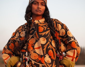 May include: A woman wearing a black embroidered jacket with orange and cream floral designs. She is wearing a beaded necklace and a headband with jewels.